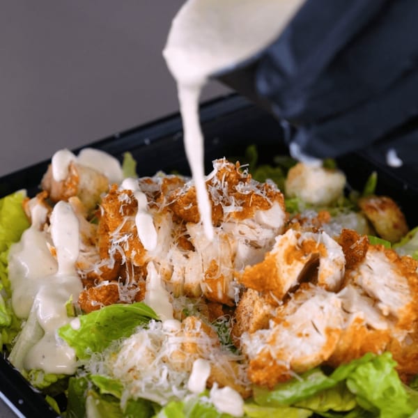 Creamy dressing being poured over a Bred Hot Chicken Caesar salad with fried chicken, lettuce, and cheese.