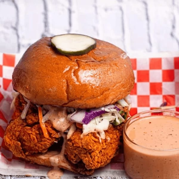 Bred Hot Chicken sandwich with fried chicken, coleslaw, pickles, and sauce, next to a cup of sauce.