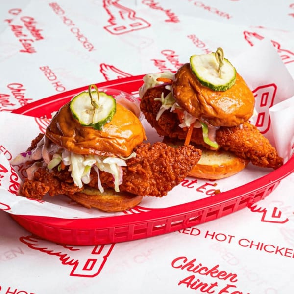 Two Bred Hot Chicken sliders with fried chicken, coleslaw, and pickles in a red basket.