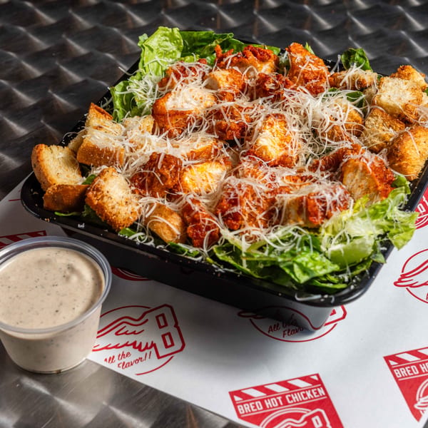 Bred Hot Chicken Caesar salad with crispy chicken, croutons, and parmesan, with a side of dressing.