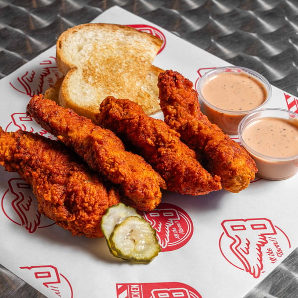Four crispy, red-orange chicken tenders with toast, pickles, and two dipping sauces on branded paper.