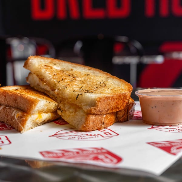 Two grilled cheese sandwiches with melted cheese and a side of dipping sauce on branded paper.