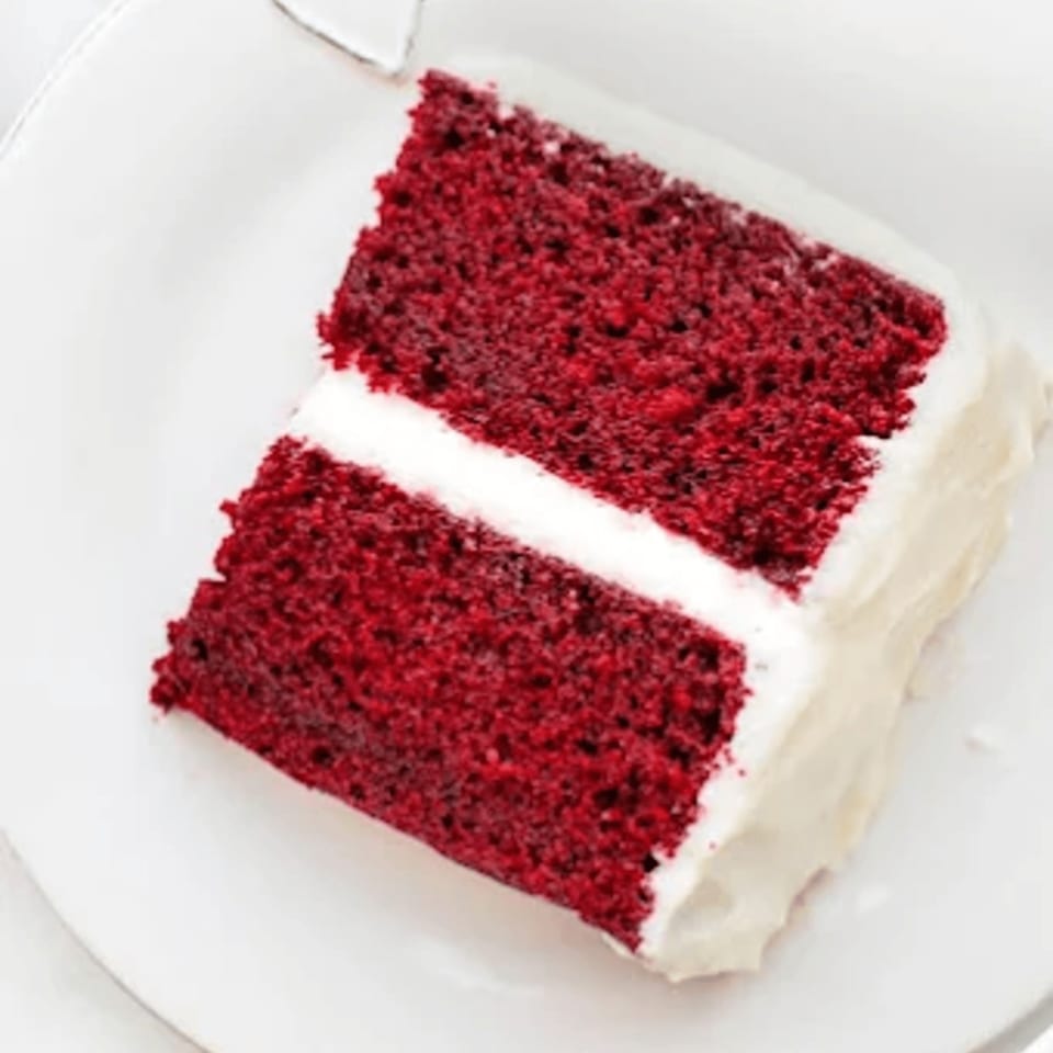 A slice of Bred Hot Chicken's red velvet cake with white frosting on a white plate.