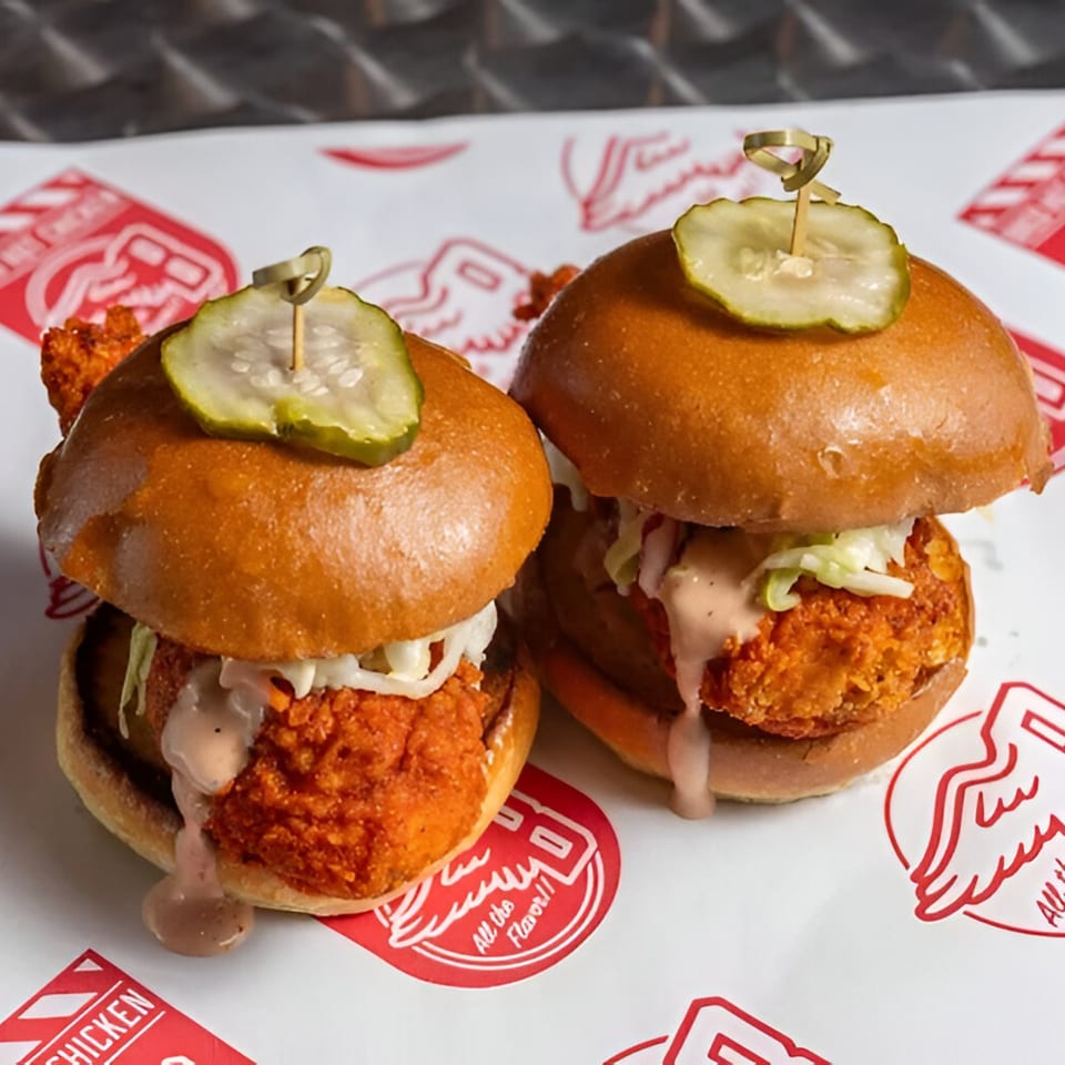 Two Bred Hot Chicken sliders with fried chicken, coleslaw, sauce, and a pickle on top.