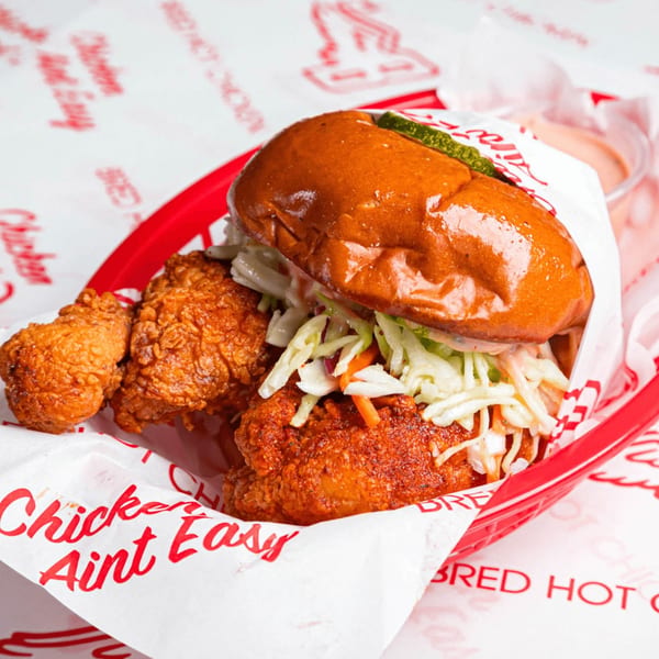 Bred Hot Chicken sandwich with coleslaw and pickles, alongside fried chicken pieces in a red basket.