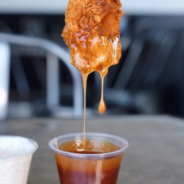 A crispy Bred Hot Chicken tender being dipped into a cup of orange sauce, with sauce dripping.